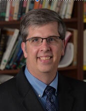 Headshot of Dan in a business suit on spectacles in front of a bookshelf